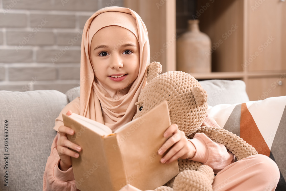 Obraz premium Little Muslim girl in hijab with teddy bear reading book at home