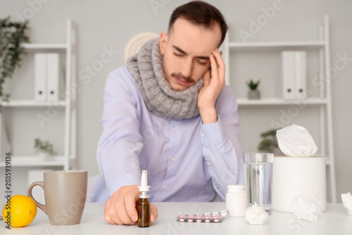 Ill businessman with scarf and nasal drops at table in office