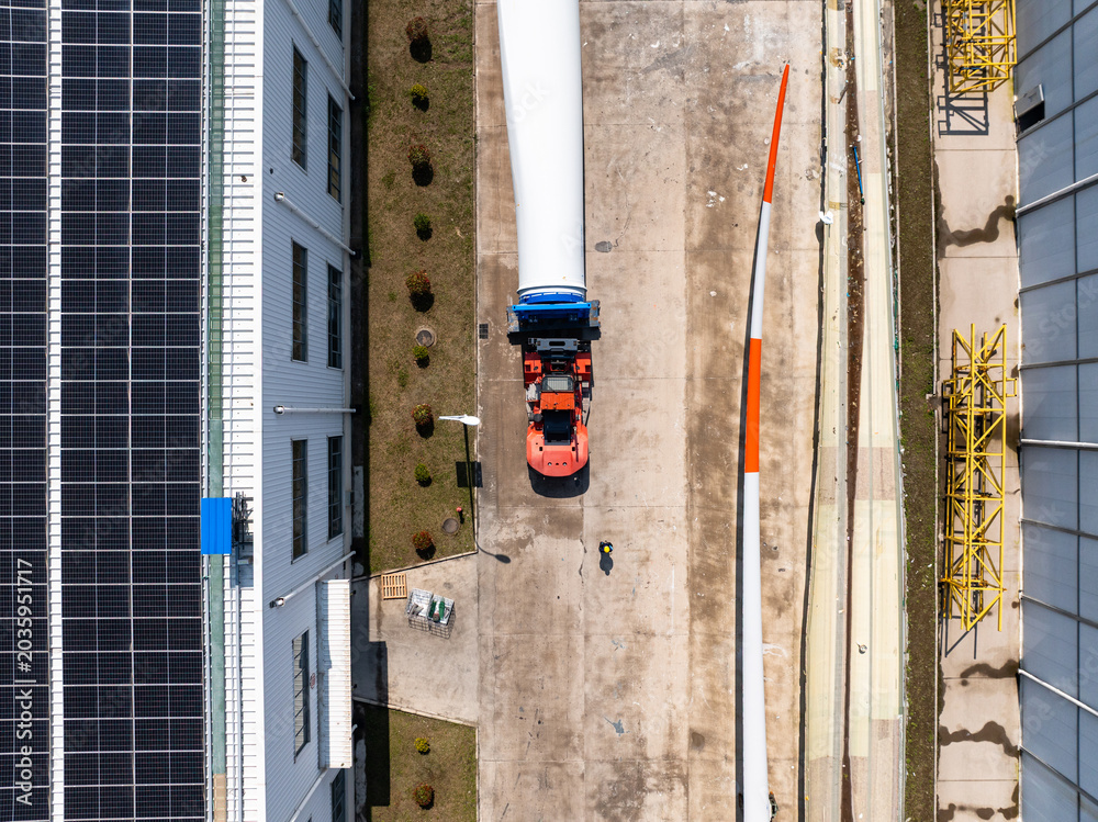 Naklejka premium Aerial top view of a specialized truck transporting a massive wind turbine blade at an industrial site