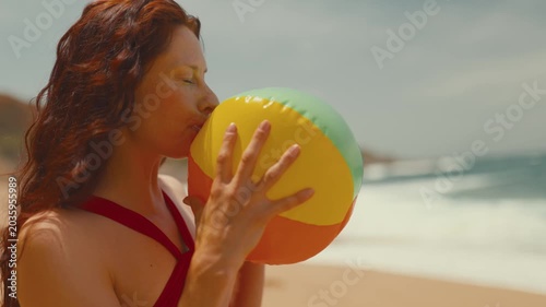 Woman with long red hair inflates a colorful beach ball at the shoreline, with gentle waves and sandy beach creating a vibrant summer atmosphere