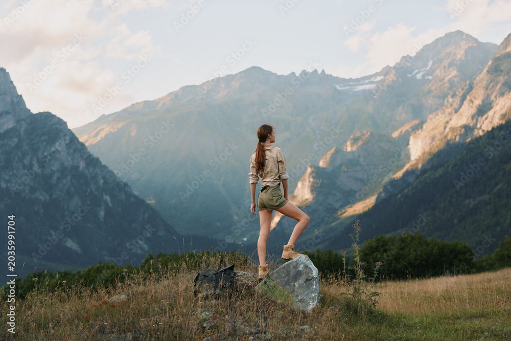 Fototapeta premium Cheerful woman outdoors, standing on a rock with distant mountains, sunny day, casual outfit, sense of adventure, natural colors, open landscape, relaxed lifestyle