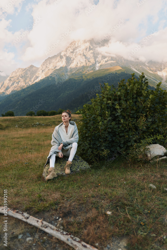 Fototapeta premium One person sits on a rock in a meadow, gazing thoughtfully at distant mountains, wrapped in a cozy cardigan for a quiet outdoor moment