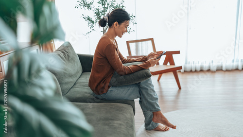 Asian woman using a smartphone while relaxing on a couch at home. Concept of modern lifestyle, communication, remote living, social media, and digital wellbeing