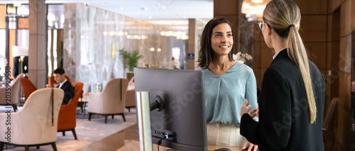 Obraz na plátne Female receptionist talking to hotel guest, providing hospitality services, explaining amenities, during check-in at front desk at inn lobby