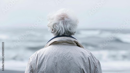 Elderly person with white hair wearing light jacket standing by ocean shore on cloudy day peaceful contemplative reflective mood