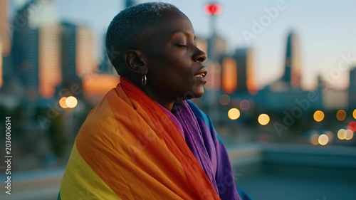 Person wrapped rainbow flag stands peacefully outdoors dusk with colorful cityscape lights celebrating LGBTQ pride equality