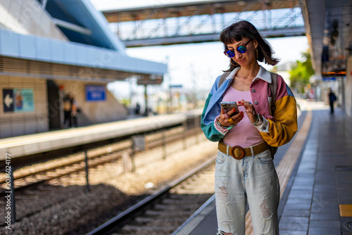 A smiling woman waits for a train and uses a smartphone at a train station. A young woman holds a smartphone and stands at a train station.