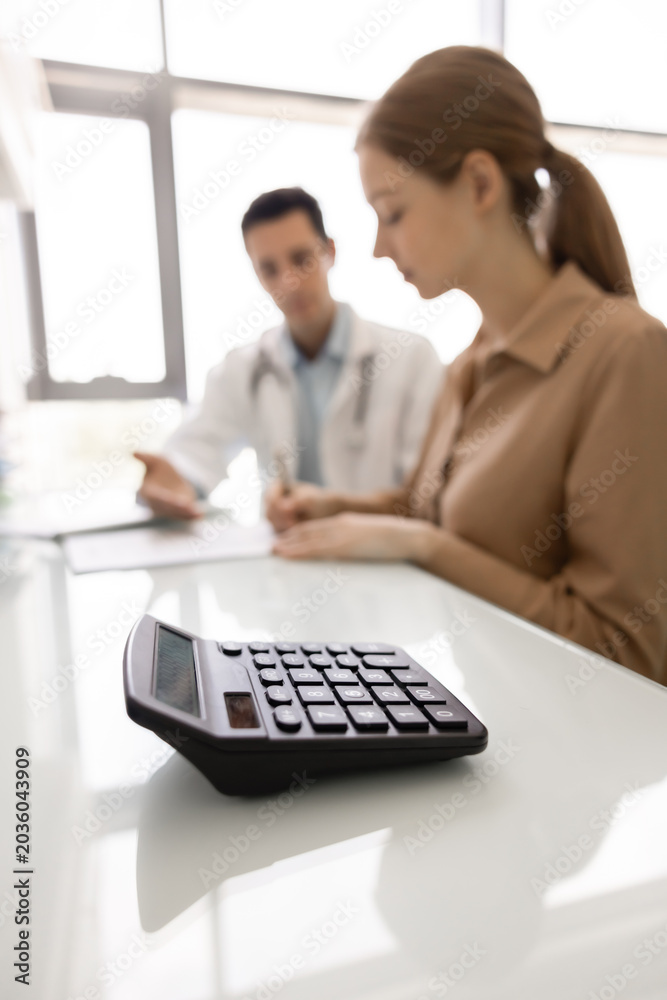 Fototapeta premium Closeup calculator lies on desk as patient consults with doctor in background, discussing charges and reviewing treatment-related documents, signing contract, get insurance coverage of private clinic