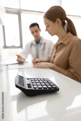 Closeup calculator lies on desk as patient consults with doctor in background, discussing charges and reviewing treatment-related documents, signing contract, get insurance coverage of private clinic