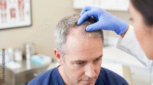 Doctor examining mature man with thinning hair and receding hairline in clinic, showing hair loss treatment, scalp checkup, dermatology care, aging, alopecia, and medical consultation.
