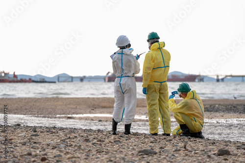 Environmental hazard inspection team monitoring coastal pollution and collecting contaminated water samples near industrial shipping zone while wearing protective hazmat equipment.