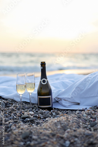 Photography Romantic evening beach picnic with a wine bottle, two filled glasses and white bedding by the sea at sunset