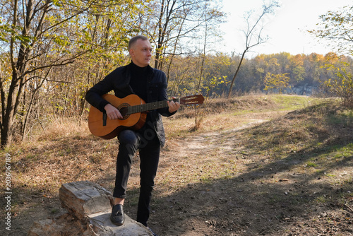 Beautiful blonde man playing acoustic guitar and singing relaxing enjoying outdoors on sunny autumn day. Artist learning and practicing playing musical instrument. Fall leaves autumnal weather 