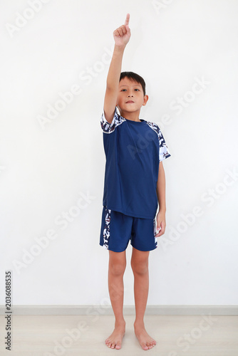 Full-length young Asian boy standing barefoot in blue and camouflage sportswear outfit, pointing one finger straight up towards the sky against white background.