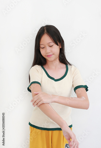 Lovely young Asian girl with long dark hair, wearing cream t-shirt with green trim, gently applying moisturizing white cream or lotion onto her arm while looking down against white background.