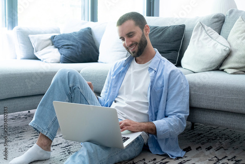 Smiling man sitting on living room floor using laptop, relaxed casual remote work at home, comfortable modern sofa and cushions in bright room, freelance, productivity, and digital lifestyle concept.