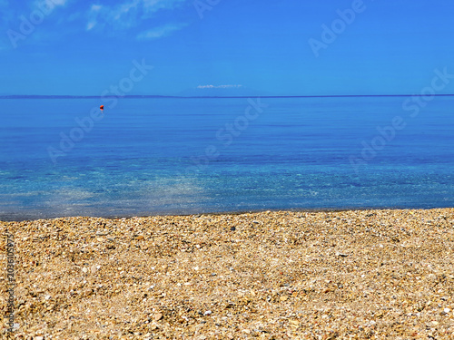 Clear Shallow Beach Shoreline With Pebbles And Blue Sky Over Calm Sea Horizon