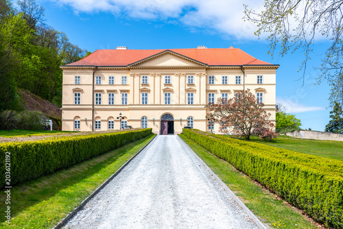 Boskovice Castle stands tall in Czechia under a bright blue sky. Visitors can see the castle's impressive facade and well-kept gardens as they walk along the gravel path leading to the entrance.
