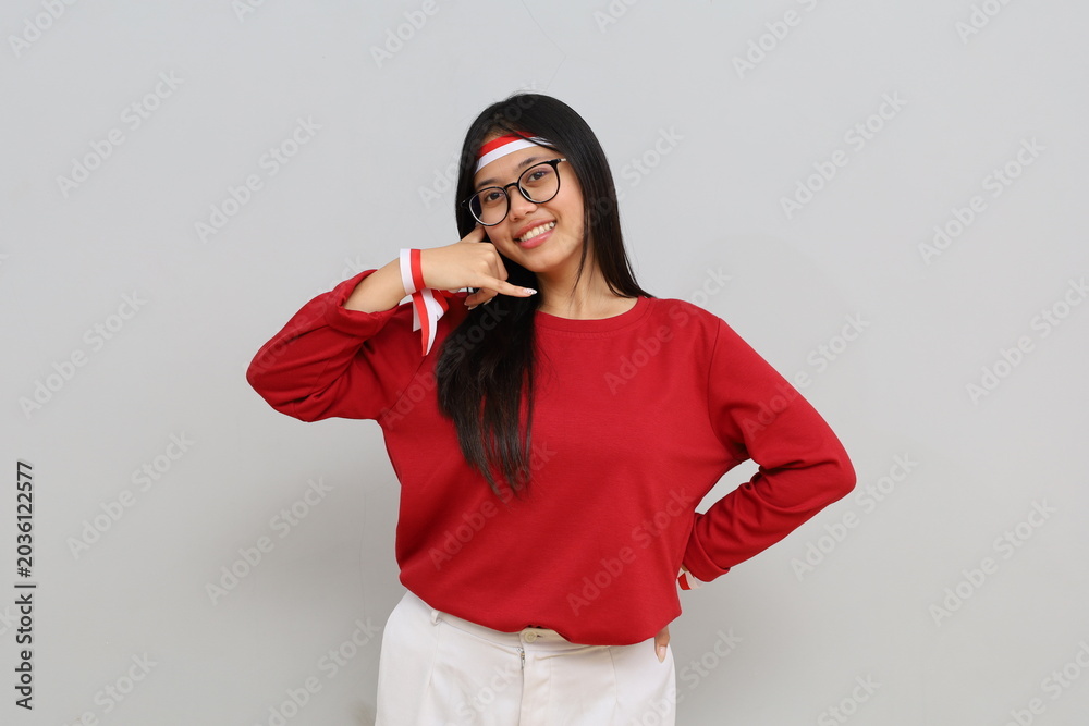 Naklejka premium Cheerful and friendly Indonesian woman with red and white ribbon celebrating Indonesian independence day while making phone hand gesture, smiling looking at camera.