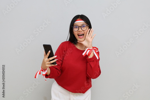 Indonesian woman with red and white ribbon to celebrate Indonesian independence day holding mobile phone while making hand gestures of announcement or promotional advertisement.