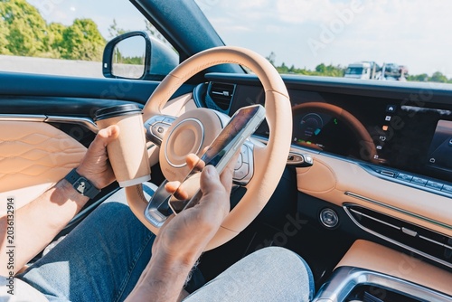 Quadro em tela Person driving luxury car while using smartphone and holding coffee cup in brigh
