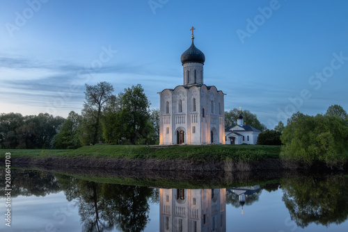 Fotografie Historic Church of the Intercession on the Nerl reflected in calm river water at sunset, famous white stone Orthodox church near Vladimir and Suzdal, Russia, scenic spring landscape with dramatic sky