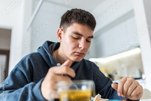 Young man eating breakfast or lunch at home kitchen. Male teenager sitting at table, eating food with spoon from bowl, having healthy meal or soup. Daily life routine, casual lifestyle concept.