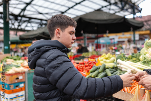 Young man paying money to vendor at outdoor farmers market. Male customer buying fresh organic vegetables and paying cash at local green marketplace. Business, trade, consumerism concept.