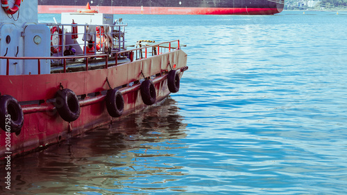 Quadro su tela Red tugboat docked in calm blue water with a large cargo ship in the background