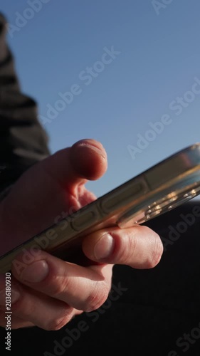A close-up view of hands typing and scrolling on a gold smartphone against a clear blue sky. Concept of digital communication, mobile networking, and modern social media lifestyle.
