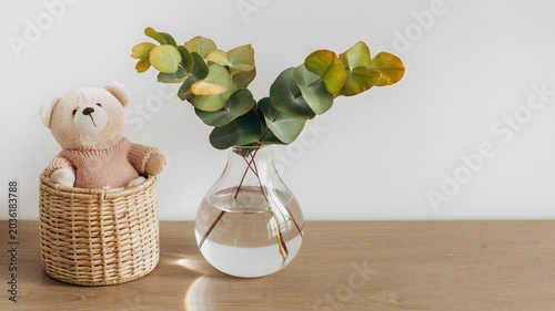Teddy Bear in Wicker Basket with Eucalyptus Branches in Glass Vase on Wooden Table