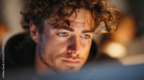 Young man focuses on work at a computer in a busy environment during evening hours while information lights glow softly