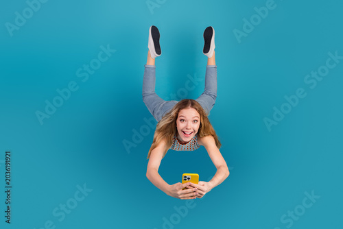 Young woman hangs upside down taking a selfie against blue background wearing casual jeans and top