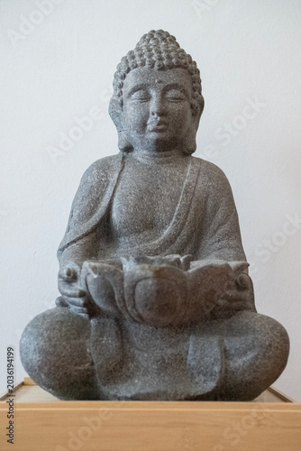 Bronze statuette depicting Buddha resting on a small wooden platform. The statuette is photographed against a light background