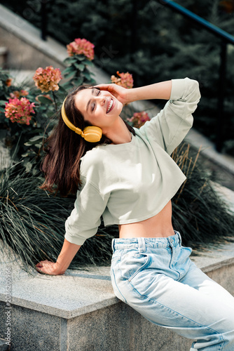 Young woman with yellow headphones smiling and relaxing outdoors on stone steps among flowers  enjoying music and sunny casual style in cropped sweatshirt and high-waisted jeans
