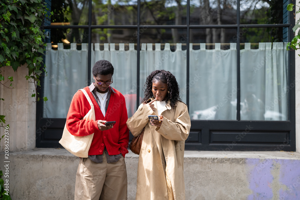 Obraz premium Thoughtful african american couple man and woman friends standing on street together browsing online on smartphones, black male and female students with unhealthy phone addiction engrossed in devices.