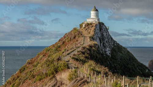 Lighthouse on coastal path under dramatic storm clouds - White coastal lighthouse viewed from a narrow path through windswept vegetation, with dark storm clouds creating a dramatic seascape atmosphere