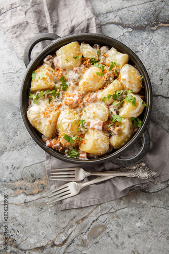 Homemade new potatoes with creamy mushroom sauce, bacon, and breadcrumbs, close-up, in a frying pan on the table. Vertical top view from above