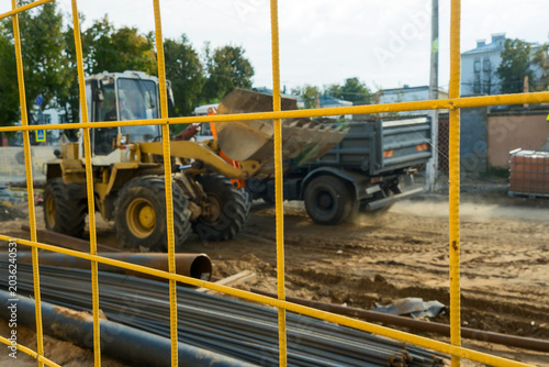 Yellow metal fence in focus with blurred construction site background. Yellow safety grid with defocused heavy machinery. Yellow wire mesh as a frame for blurry city renovation and development.