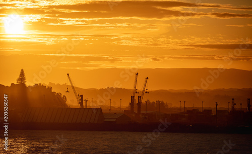 Dramatic golden sunset over an industrial harbour with silhouetted cranes, water in the foreground and hazy hills in the distance. Atmospheric maritime, shipping and industrial landscape photography