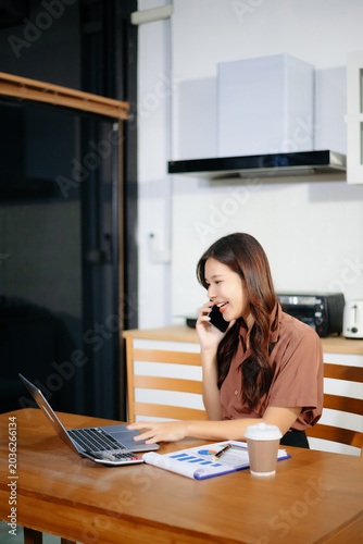 Asian woman talking on her phone while working remotely on a laptop at a kitchen table. Ideal for work from home and freelance concepts.
