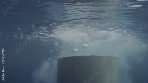 Underwater scene showing a cylindrical object submerged in water, creating bubbles and turbulence as air escapes, with rippling surface above and clear blue water surrounding
