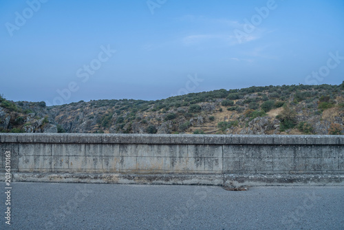Tableau sur toile A weathered stone bridge parapet overlooks a paved road, with rocky hills and scattered vegetation under a cloudy blue sky