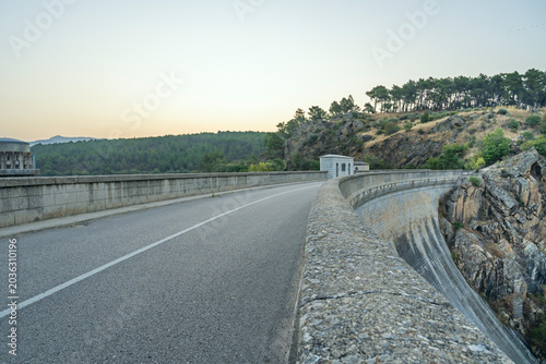 Tableau sur toile A winding road of dark asphalt cuts its way between two enormous turquoise bodies of water, surrounded by hills covered in holm oaks and dry scrubland that reflect the light of the setting sun