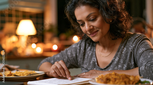 Middle-aged South Asian woman at modest kitchen table tracing handwritten food journal entries with fingertip, half-eaten comfort meal pushed aside, late evening lamp glow, recognizing emotional hun