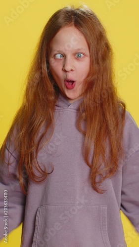 Cheerful funny bully young cute girl showing tongue making faces at camera, fooling around, joking, aping with silly face, teasing. Preteen redhead child kid on yellow background. Little children