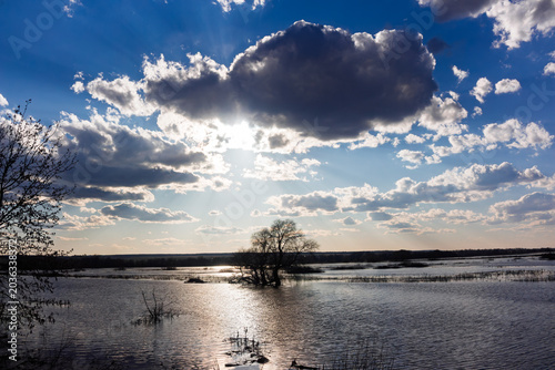 Fotografie Spring flood inundates fields near Ogublyanka Lake, featuring dramatic sunbeams breaking through heavy clouds above a solitary tree reflecting in the rippling water