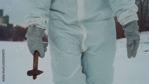 Hazmat suited scientist walking across snow holding rusted crucifix, gloved hand close up, abandoned riverbank and distant industrial skyline, cold pale light and eerie silence, slow deliberate steps