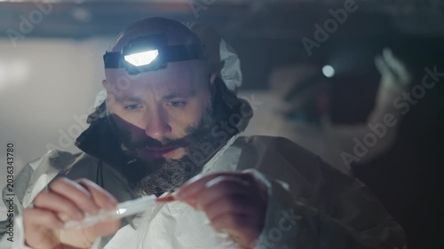 White technician under lamp carefully unwraps and tests suspected nuclear sample, focused gloved hands, respirator visible, monitoring and logging procedure in dim bunker lab, atmosphere of urgency