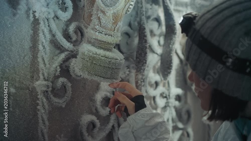 Young explorer tracing frost on ornate gate headlamp beam reveals icy filigree, gloved fingers gently touch metal pattern, winter hush surrounds abandoned house facade, careful sample collection